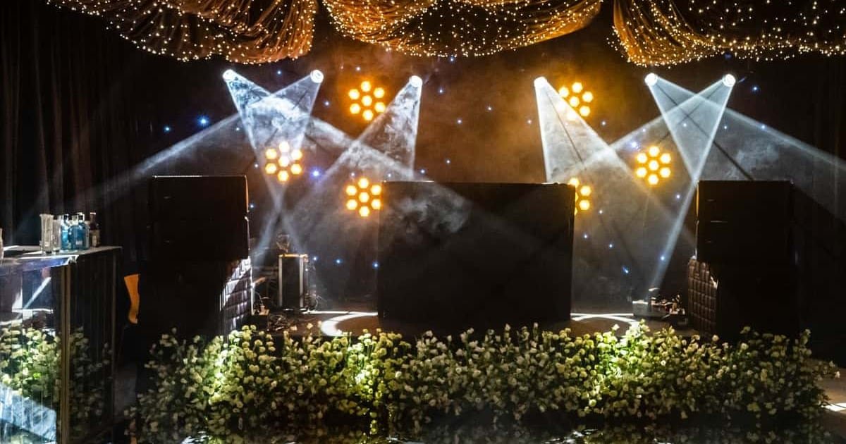 Beams of warm stage light cutting through haze above a bank of white flowers, under a canopy of fairy lights
